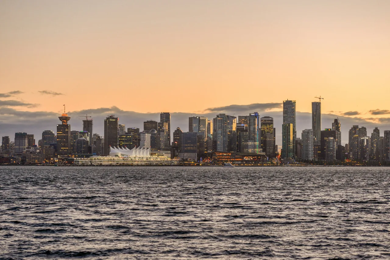 Vancouvers skyline og dens hvide bygning med sejlformet tag ved vandkanten, der gløder i solnedgangen, set over vandet